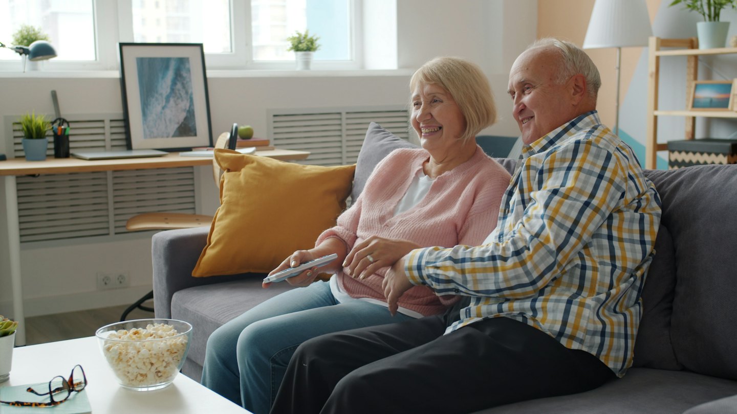 An older couple smiles on a sofa while holding a TV remote.